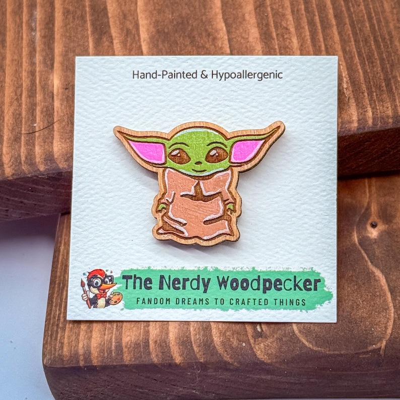 A hand-painted wooden pin depicting the character Baby Yoda from the Star Wars franchise, with the text "Hand-painted & Hypoallergenic" and "The Nerdy Woodpecker FANDOM DREAMS TO CRAFTED THINGS" printed on a white background.