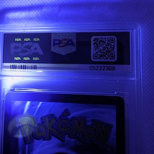 K&ouml;nnte beinhalten: Eine bewertete Sammelkarte in einem transparenten Kunststoffhalter. Der Halter zeigt das PSA-Logo und einen QR-Code. Der untere Teil der Karte zeigt das Wort "Pok&eacute;mon" in Gelb und Orange.