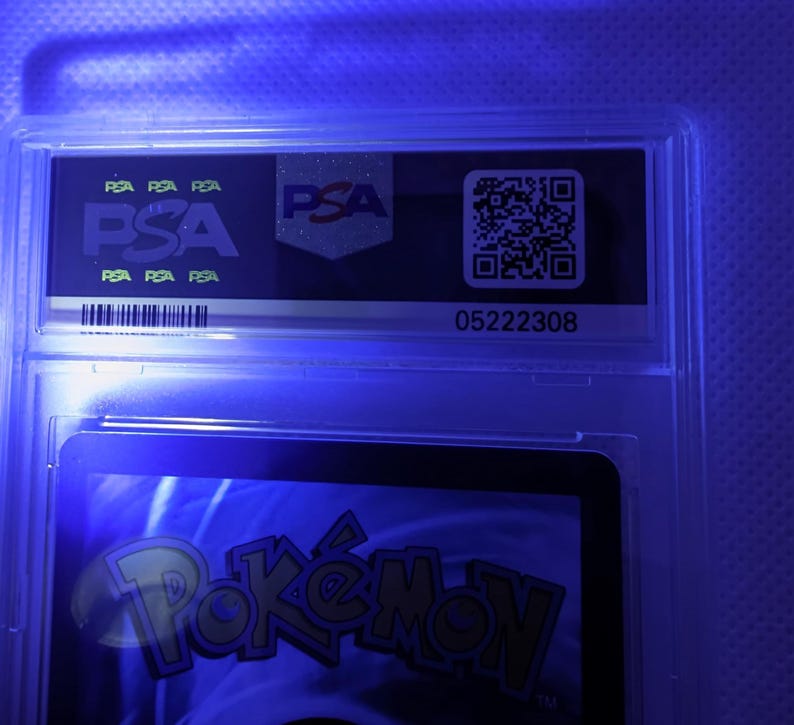 K&ouml;nnte beinhalten: Eine bewertete Sammelkarte in einem transparenten Kunststoffhalter. Der Halter zeigt das PSA-Logo und einen QR-Code. Der untere Teil der Karte zeigt das Wort "Pok&eacute;mon" in Gelb und Orange.