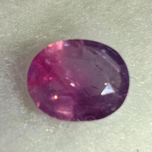 May include: A close-up of a purple and pink oval gemstone. The gemstone has a smooth, polished surface and is set against a white background.