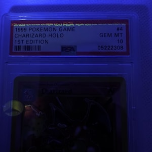 K&ouml;nnte beinhalten: Eine bewertete Pok&eacute;mon-Karte in einer Schutzh&uuml;lle. Die Karte ist eine 1999 Charizard-Holo 1st Edition, bewertet mit GEM MT 10 von PSA. Die Kartendetails sind auf einem rot-wei&szlig;en Etikett aufgedruckt.