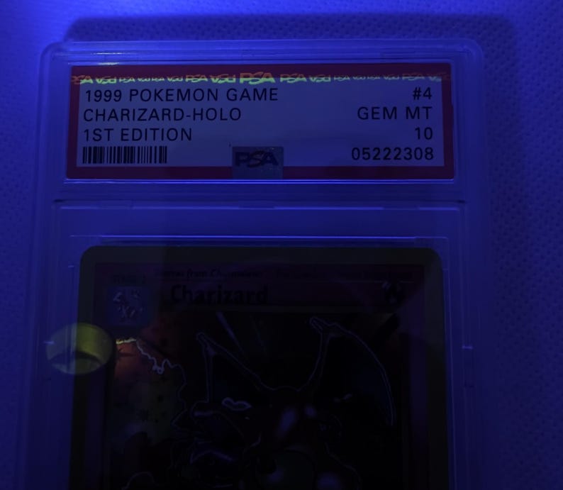 K&ouml;nnte beinhalten: Eine bewertete Pok&eacute;mon-Karte in einer Schutzh&uuml;lle. Die Karte ist eine 1999 Charizard-Holo 1st Edition, bewertet mit GEM MT 10 von PSA. Die Kartendetails sind auf einem rot-wei&szlig;en Etikett aufgedruckt.
