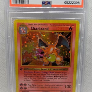 K&ouml;nnte beinhalten: Eine bewertete 1999 Pok&eacute;mon Charizard-Holo 1st Edition Karte. Die Karte zeigt eine lebendige Illustration von Charizard, einer orange-gelben drachenartigen Kreatur. Die Karte ist in einem Schutzhalter mit einer PSA-Note von 10 eingeschlossen.