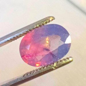 May include: A close-up of a pink and purple oval gemstone held by tweezers. The gemstone has a slight shimmer and is cut into a faceted shape.