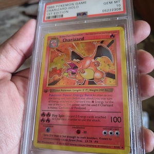 K&ouml;nnte beinhalten: Eine bewertete 1999 Pok&eacute;mon Game Charizard Holo 1st Edition Karte. Die Karte zeigt eine Charizard-Illustration mit einem rot-orangen Farbschema. Die Karte ist in einem Schutzhalter mit einer PSA-Note von 10 eingeschlossen.
