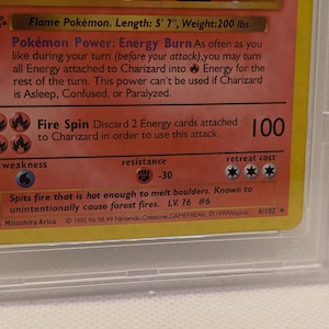 K&ouml;nnte beinhalten: Nahaufnahme einer Charizard Pok&eacute;mon-Karte, in einer Schutzh&uuml;lle. Die Karte enth&auml;lt Text, der die F&auml;higkeiten des Charakters beschreibt, darunter "Fire Spin" und "Energy Burn". Die Karte ist gelb und rot mit einem Design zum Thema Feuer. Die Karte ist 170 cm lang.