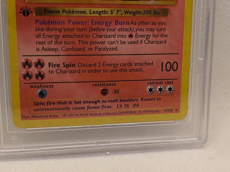 K&ouml;nnte beinhalten: Nahaufnahme einer Charizard Pok&eacute;mon-Karte, in einer Schutzh&uuml;lle. Die Karte enth&auml;lt Text, der die F&auml;higkeiten des Charakters beschreibt, darunter "Fire Spin" und "Energy Burn". Die Karte ist gelb und rot mit einem Design zum Thema Feuer. Die Karte ist 170 cm lang.