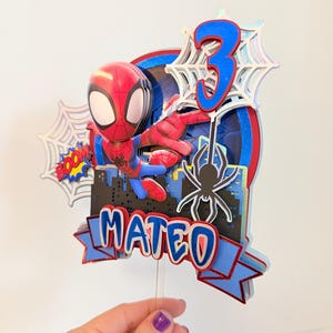 May include: A Spiderman-themed cake topper with a 3D Spiderman figure, a blue number 3, and the name "Mateo". The design incorporates a cityscape silhouette, spiderwebs, and the word "BOOM!" in a comic book style.