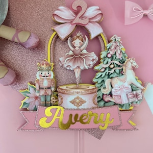 Op de afbeelding: Een roze en gouden ballerina-thema taarttopper met de naam "Avery". De topper heeft een ballerina, een notenkraker, een kerstboom met balletschoenen en een speelgoedpaard. Bovenop zit een grote roze strik met het nummer "2".