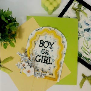 Op de afbeelding: Een taarttopper voor een gender reveal, met de tekst "BOY OR GIRL". De topper heeft een geel en wit geschulpte lijst met bloemendetails. Een groene vierkant en gele envelop staan op de achtergrond, samen met een groene kaars en een ingelijste foto.