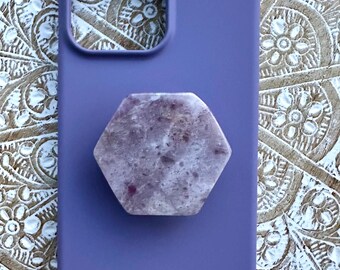 May include: A periwinkle phone case with a hexagonal, speckled, light purple phone grip. The phone case has a smooth, matte finish. The phone grip is made of a stone-like material.