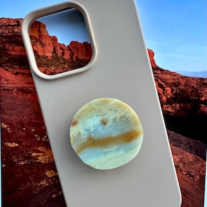 May include: A light beige phone case with a circular, multi-colored phone grip. The grip features shades of blue, tan, and white. The phone case has a cutout for the camera. The background shows a landscape with red rock formations.