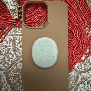 May include: A tan phone case with a light green circular grip attached. The case has a rectangular camera cutout. The background includes a red beaded necklace and a decorative skull.