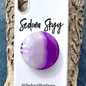 May include: White phone case with a purple and white agate stone pop socket. The case has the text "Sedona Skyy" and "@SedonaSkyyGems" printed on it.