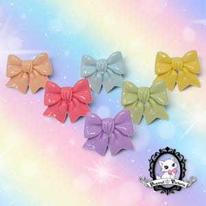 May include: A collection of six glossy, solid-colored bow-shaped charms in peach, light blue, yellow, hot pink, light green, and lavender. Each bow features a central knot detail and smooth, rounded edges. A logo with a cat and the text Dearest Victoria.