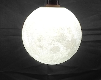 May include: A white moon-shaped lamp with a textured surface sits on a wooden tripod stand. The lamp is lit and casts a soft glow.