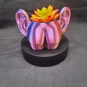 Op de afbeelding: Een 3D-geprinte plantenbak in een verloop van roze, paars en blauw, gevormd als een gestileerde figuur. De plantenbak bevat een kunstmatige vetplant met gele, oranje en groene bladeren. De plantenbak staat op een zwarte cirkelvormige basis.