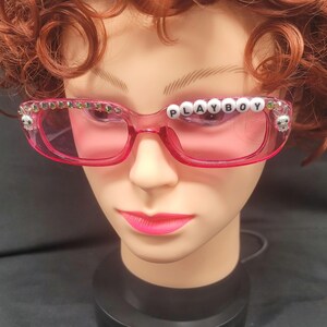 May include: Pink-tinted sunglasses with the word "PLAYBOY" spelled out in white letter beads across the top. The frames are adorned with small skulls and green gems. The sunglasses are on a mannequin head with red hair.