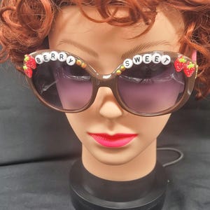 Puede incluir: Gafas de sol marrones de ojo de gato con las palabras "BERRY SWEET" escritas en letras blancas con adornos de fresas rojas y verdes.