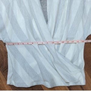 May include: White and gray striped wrap top with a V-neck design. A measuring tape is laid across the garment, showing a measurement of approximately 17 inches. The top is made of a soft, lightweight fabric.