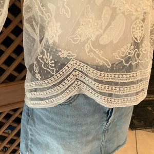 Pu&ograve; includere: Top in pizzo bianco con intricati motivi floreali e geometrici. Il top presenta un tessuto delicato e trasparente con ricami dettagliati e un orlo smerlato. Il top &egrave; abbinato a una gonna di jeans blu chiaro.