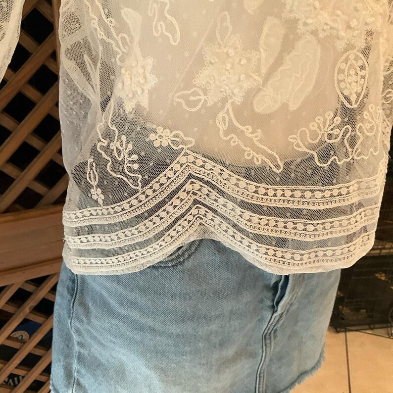 Pu&ograve; includere: Top in pizzo bianco con intricati motivi floreali e geometrici. Il top presenta un tessuto delicato e trasparente con ricami dettagliati e un orlo smerlato. Il top &egrave; abbinato a una gonna di jeans blu chiaro.