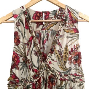 May include: Cream sleeveless dress with a colourful floral pattern, including red, yellow, and green botanical designs. The dress has a ruffled trim around the neckline and a V-neck. It is displayed on a wooden hanger.