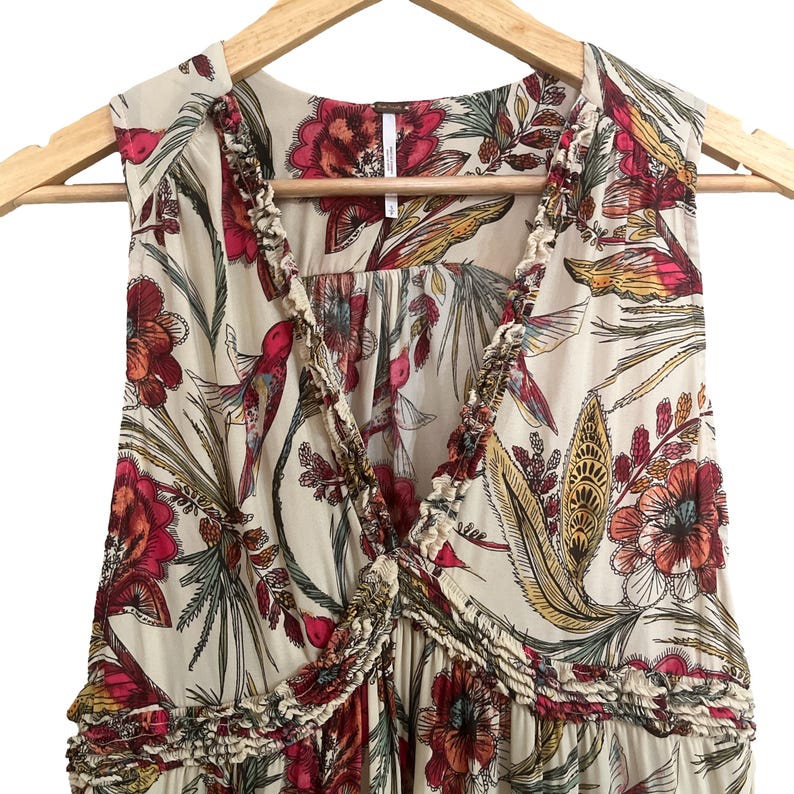 May include: Cream sleeveless dress with a colourful floral pattern, including red, yellow, and green botanical designs. The dress has a ruffled trim around the neckline and a V-neck. It is displayed on a wooden hanger.