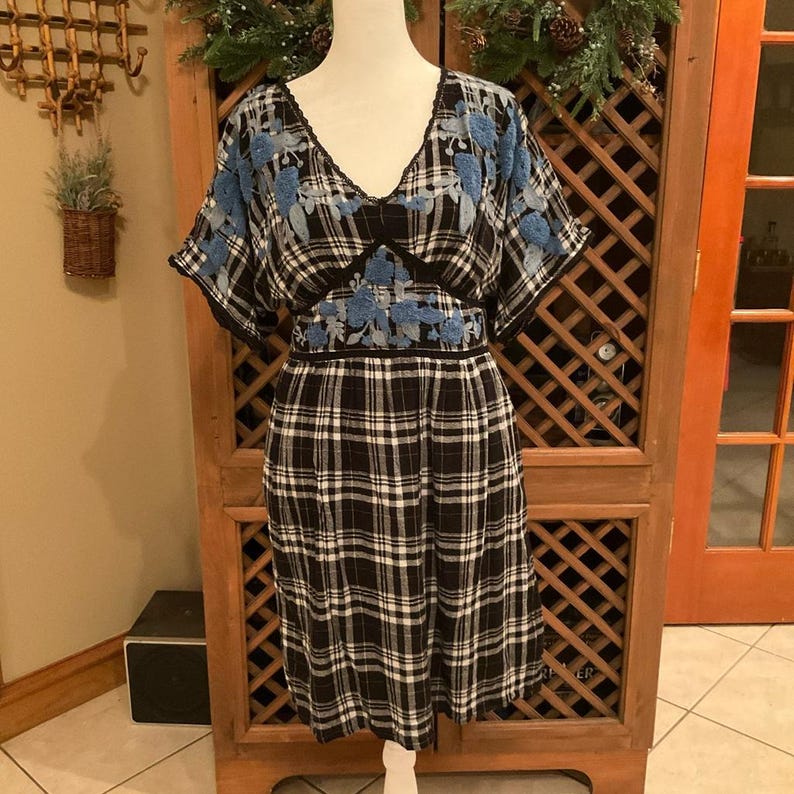 May include: A black and white plaid dress with blue floral embroidery on the sleeves and bodice. The dress has a V-neck and short sleeves. The dress is displayed on a mannequin.
