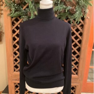 May include: A navy blue turtleneck sweater displayed on a white mannequin. The long-sleeved sweater has a ribbed hem and cuffs. The background includes a wooden door and a decorative wreath with pinecones.