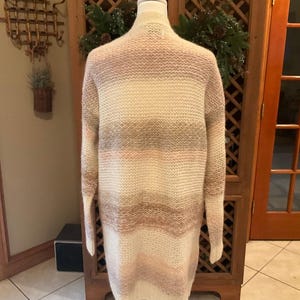 May include: A long, open-front cardigan in a striped pattern of cream, light pink, and taupe. The sweater is displayed on a white mannequin, showcasing its length and relaxed fit. The knitwear is soft and cozy, perfect for layering.
