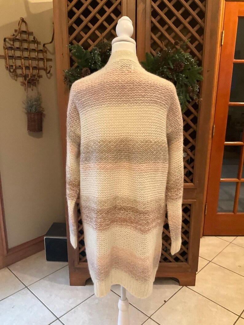 May include: A long, open-front cardigan in a striped pattern of cream, light pink, and taupe. The sweater is displayed on a white mannequin, showcasing its length and relaxed fit. The knitwear is soft and cozy, perfect for layering.
