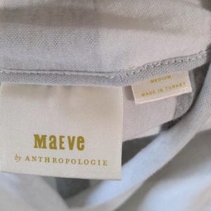 May include: Close-up of a clothing tag with the brand name "Maeve" in gold lettering, with "by ANTHROPOLOGIE" below. The tag also indicates the size "MEDIUM" and "MADE IN TURKEY". The fabric is a light gray and white striped pattern.