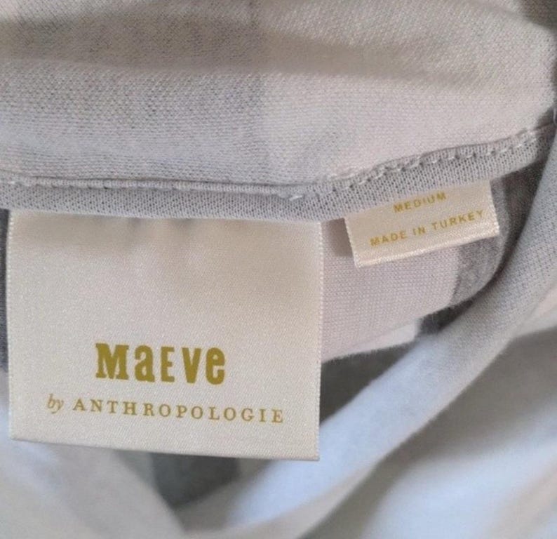 May include: Close-up of a clothing tag with the brand name "Maeve" in gold lettering, with "by ANTHROPOLOGIE" below. The tag also indicates the size "MEDIUM" and "MADE IN TURKEY". The fabric is a light gray and white striped pattern.