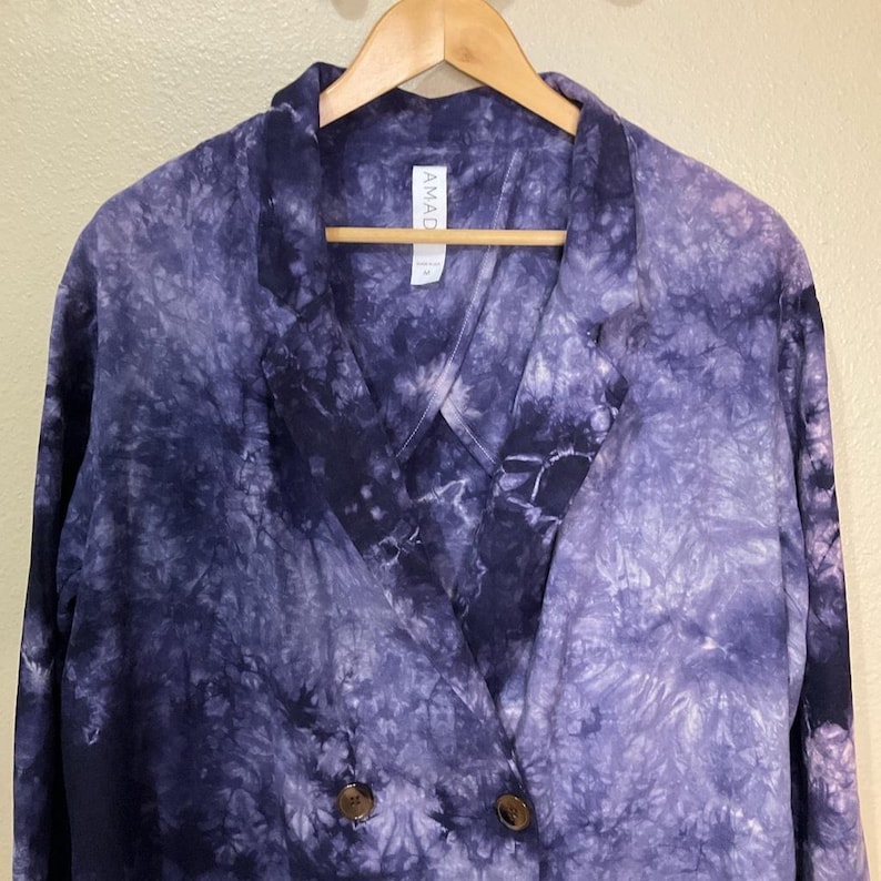 May include: A tie-dye blazer in shades of indigo and lavender, featuring a double-breasted design with two brown buttons. The blazer is hanging on a wooden hanger. The tag reads "AMAD".