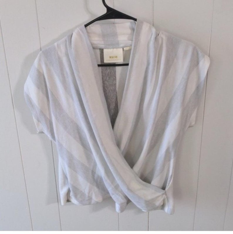 May include: A white and gray striped wrap top with short sleeves. The top is hanging on a black hanger against a white wall. The top has a draped design and a tag that reads "MEIVE".