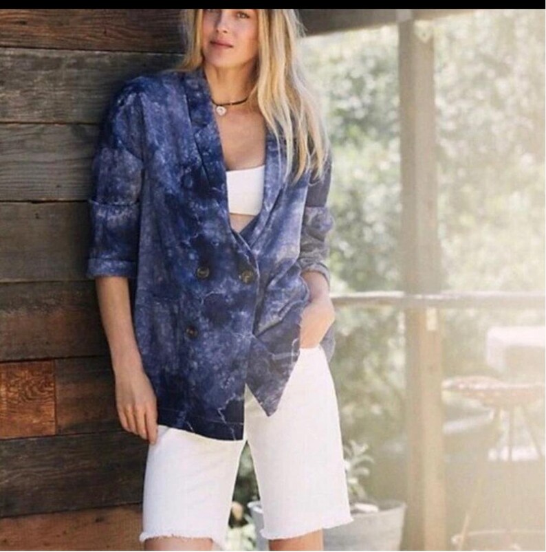 May include: A double-breasted blazer in a blue tie-dye pattern. The blazer has a notched lapel and button closure. Paired with white denim shorts with a frayed hem. The outfit is casual and stylish.