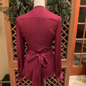 Puede incluir: Blusa burdeos con mangas largas y lazo envolvente en la cintura. La blusa se exhibe en un maniqu&iacute;, mostrando su ajuste y dise&ntilde;o a medida. Los pu&ntilde;os tienen cierre de bot&oacute;n. El fondo incluye un armario de madera y una corona decorativa.