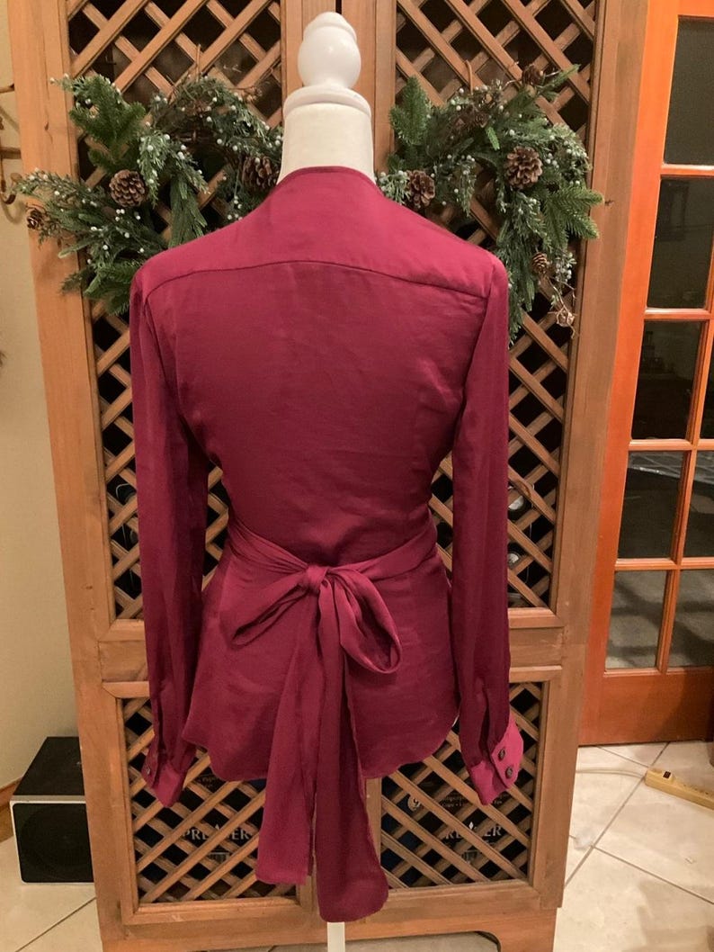 Puede incluir: Blusa burdeos con mangas largas y lazo envolvente en la cintura. La blusa se exhibe en un maniqu&iacute;, mostrando su ajuste y dise&ntilde;o a medida. Los pu&ntilde;os tienen cierre de bot&oacute;n. El fondo incluye un armario de madera y una corona decorativa.