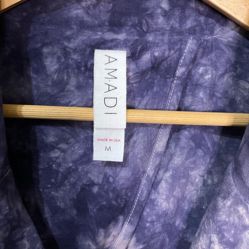 May include: Close-up of a tie-dye shirt in shades of purple and blue, hanging on a wooden hanger. The shirt has a tag that reads "AMADI" and "MADE IN USA", with a size "M". The fabric has a textured appearance.