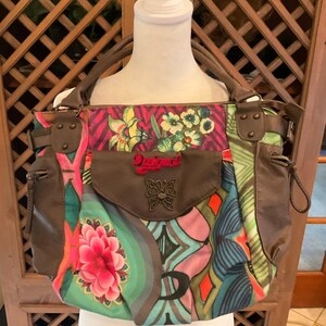 May include: A colorful handbag with a floral and geometric pattern. The bag features a brown flap with a butterfly emblem and the word "Desigual" in red. It has brown handles and side pockets.