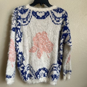 May include: White fuzzy sweater with a blue and white geometric pattern and a pink floral design.