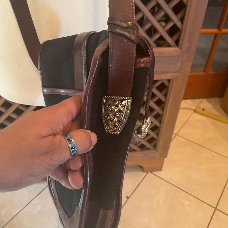 May include: Black crossbody bag with brown leather trim and strap. The bag features a decorative silver-tone buckle and a woven texture on the front. The bag is held by a person.