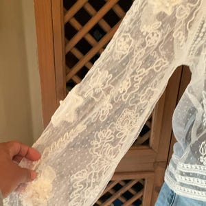Pu&ograve; includere: Primo piano di una manica in pizzo color crema. La manica presenta intricati motivi floreali e a spirale, con una texture delicata e trasparente. Il design include piccole applicazioni floreali in rilievo. La manica fa parte di un capo.