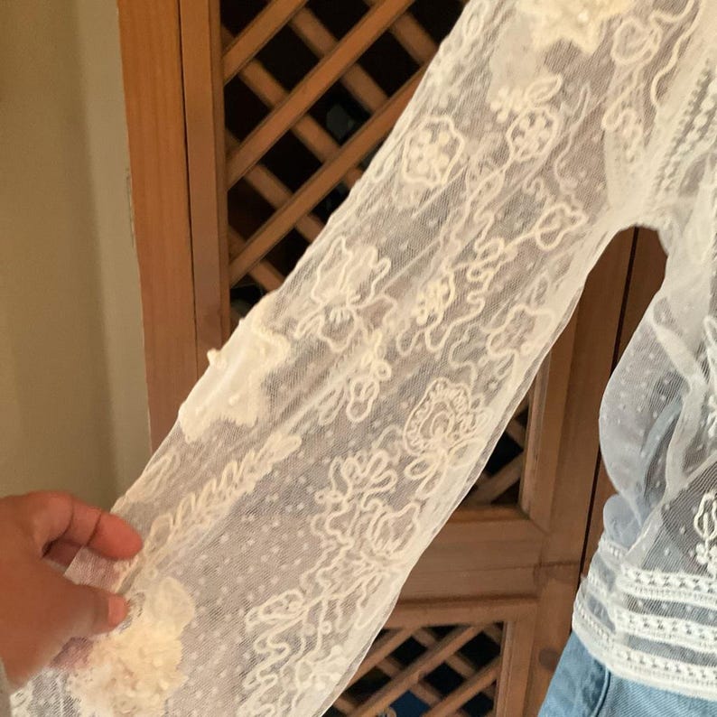 Pu&ograve; includere: Primo piano di una manica in pizzo color crema. La manica presenta intricati motivi floreali e a spirale, con una texture delicata e trasparente. Il design include piccole applicazioni floreali in rilievo. La manica fa parte di un capo.