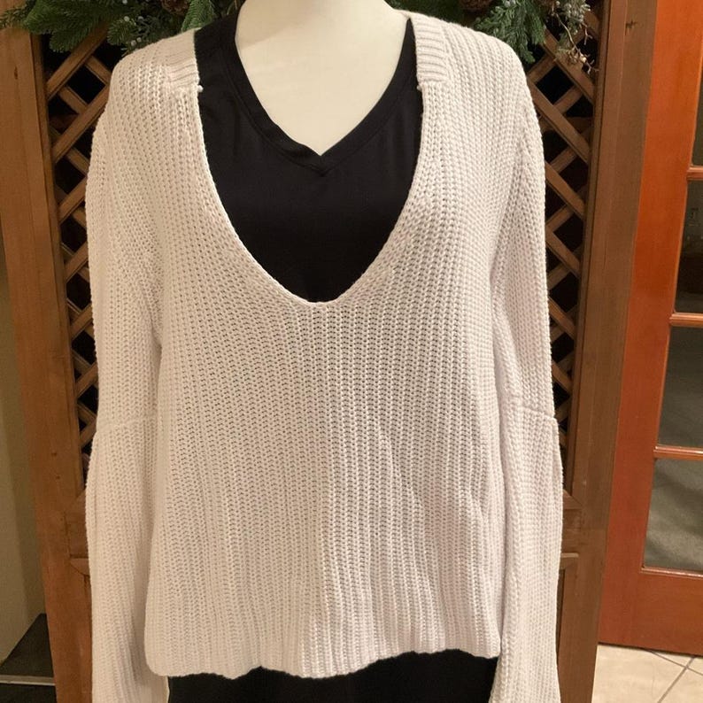 May include: A white, open-knit sweater with a deep V-neck. The sweater has long sleeves and a loose fit. A black V-neck shirt is visible underneath. The sweater is displayed on a mannequin.