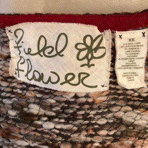 May include: Close-up of a fabric tag from a garment. The tag is white with the words "Field Flower" in a handwritten font. A smaller tag lists the fabric composition, including wool, acrylic, and cashmere. The garment is made in China.