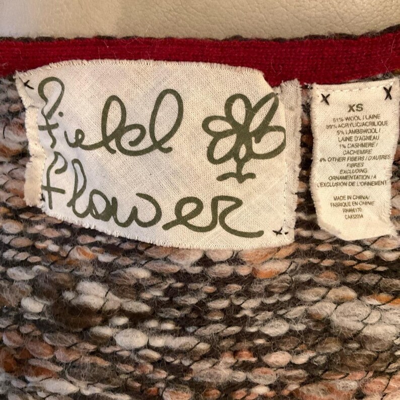 May include: Close-up of a fabric tag from a garment. The tag is white with the words "Field Flower" in a handwritten font. A smaller tag lists the fabric composition, including wool, acrylic, and cashmere. The garment is made in China.
