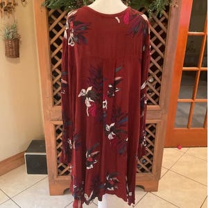 May include: A long-sleeved, burgundy dress with a floral pattern in white, blue, and pink. The dress is displayed on a white mannequin. The dress has a loose fit and a round neckline. The background includes a wooden cabinet and a decorative wreath.