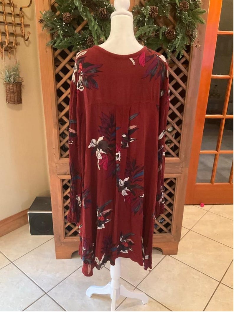 May include: A long-sleeved, burgundy dress with a floral pattern in white, blue, and pink. The dress is displayed on a white mannequin. The dress has a loose fit and a round neckline. The background includes a wooden cabinet and a decorative wreath.